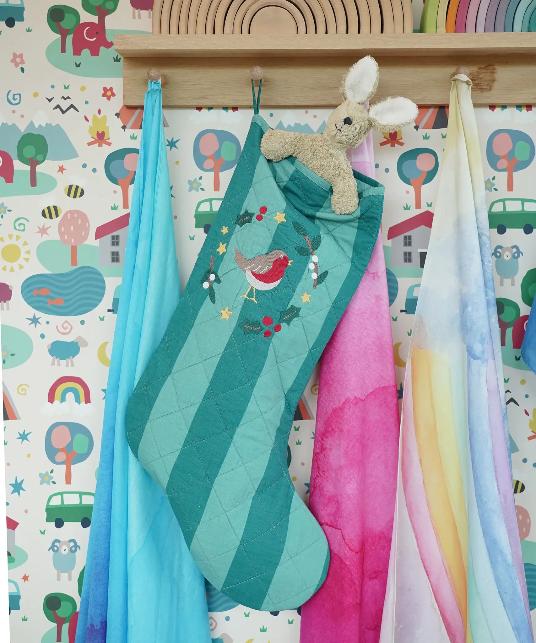 Frugi organic cotton Christmas stocking. Striped teal quilted style stocking with a robin wreath embroidery detail and hanging loop hung up in the Babipur playroom with a Senger rabbit soft toy inside.