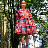 Girl wearing a Frugi festive check red, blue, and yellow plaid long-sleeve dress with ruffled shoulders and brown boots.