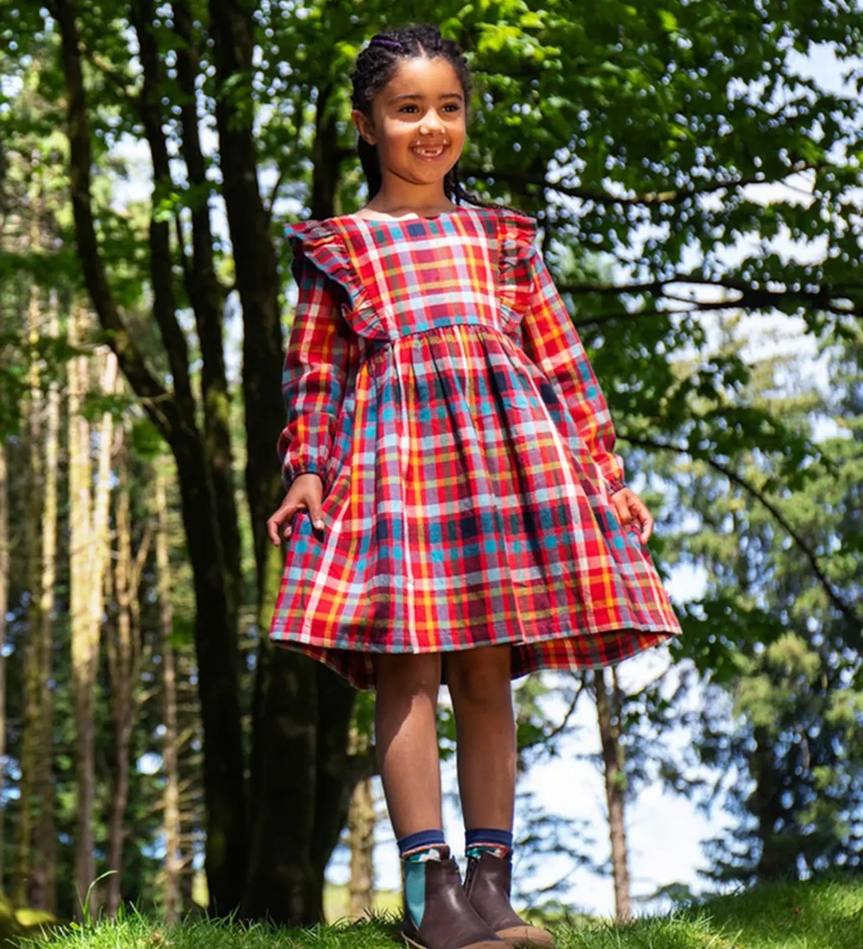 Girl wearing a Frugi festive check red, blue, and yellow plaid long-sleeve dress with ruffled shoulders and brown boots.