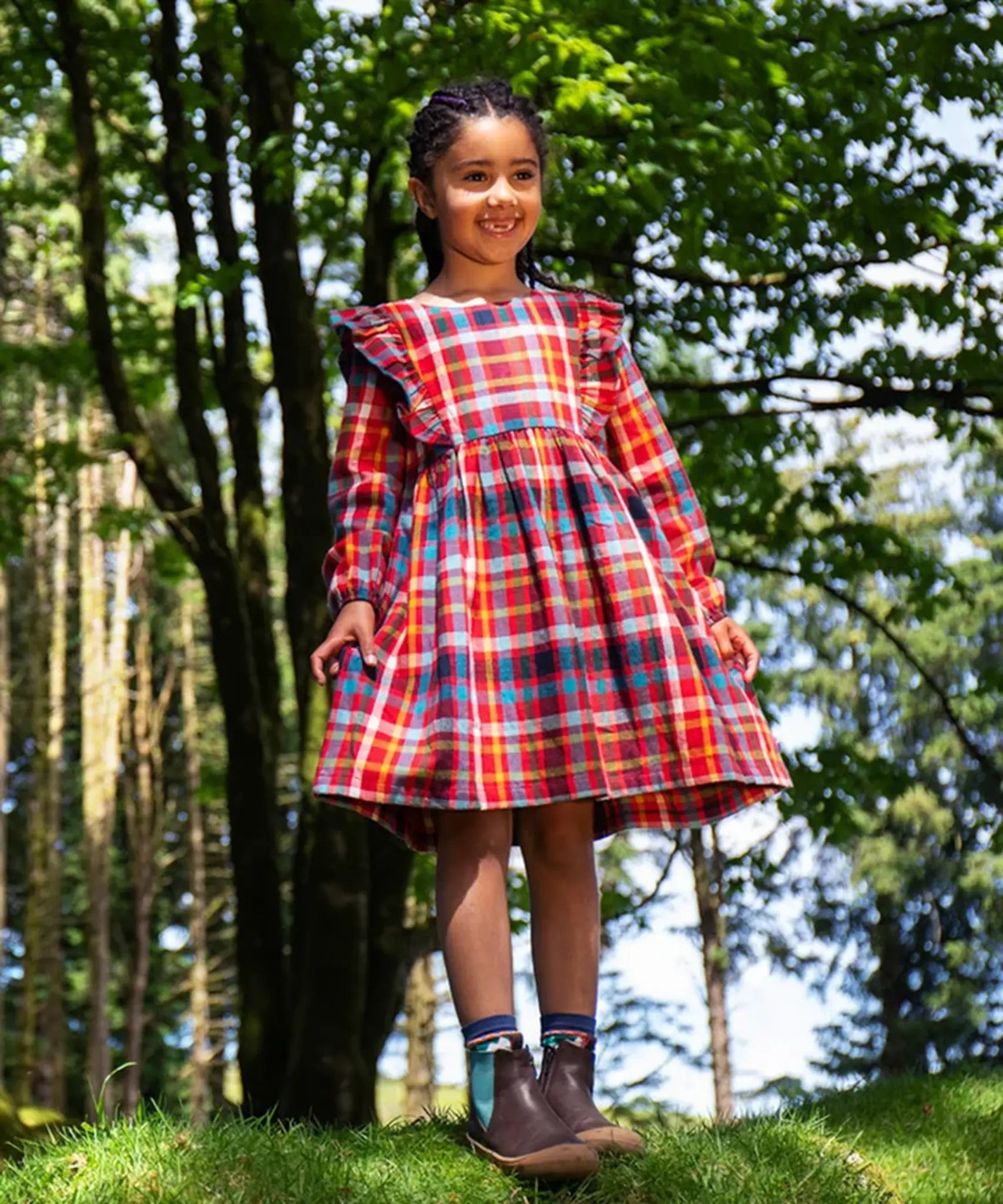 Girl wearing a Frugi festive check red, blue, and yellow plaid long-sleeve dress with ruffled shoulders and brown boots.