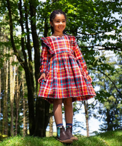 Girl wearing a Frugi festive check red, blue, and yellow plaid long-sleeve dress with ruffled shoulders and brown boots.