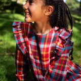 Close-up of a child wearing a Frugi festive check long-sleeve plaid dress outdoors, showing the ruffled shoulder details on the front. The dress features a busy multicolor plaid pattern with red, blue, yellow, green, and white.