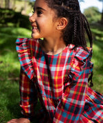 Close-up of a child wearing a Frugi festive check long-sleeve plaid dress outdoors, showing the ruffled shoulder details on the front. The dress features a busy multicolor plaid pattern with red, blue, yellow, green, and white.