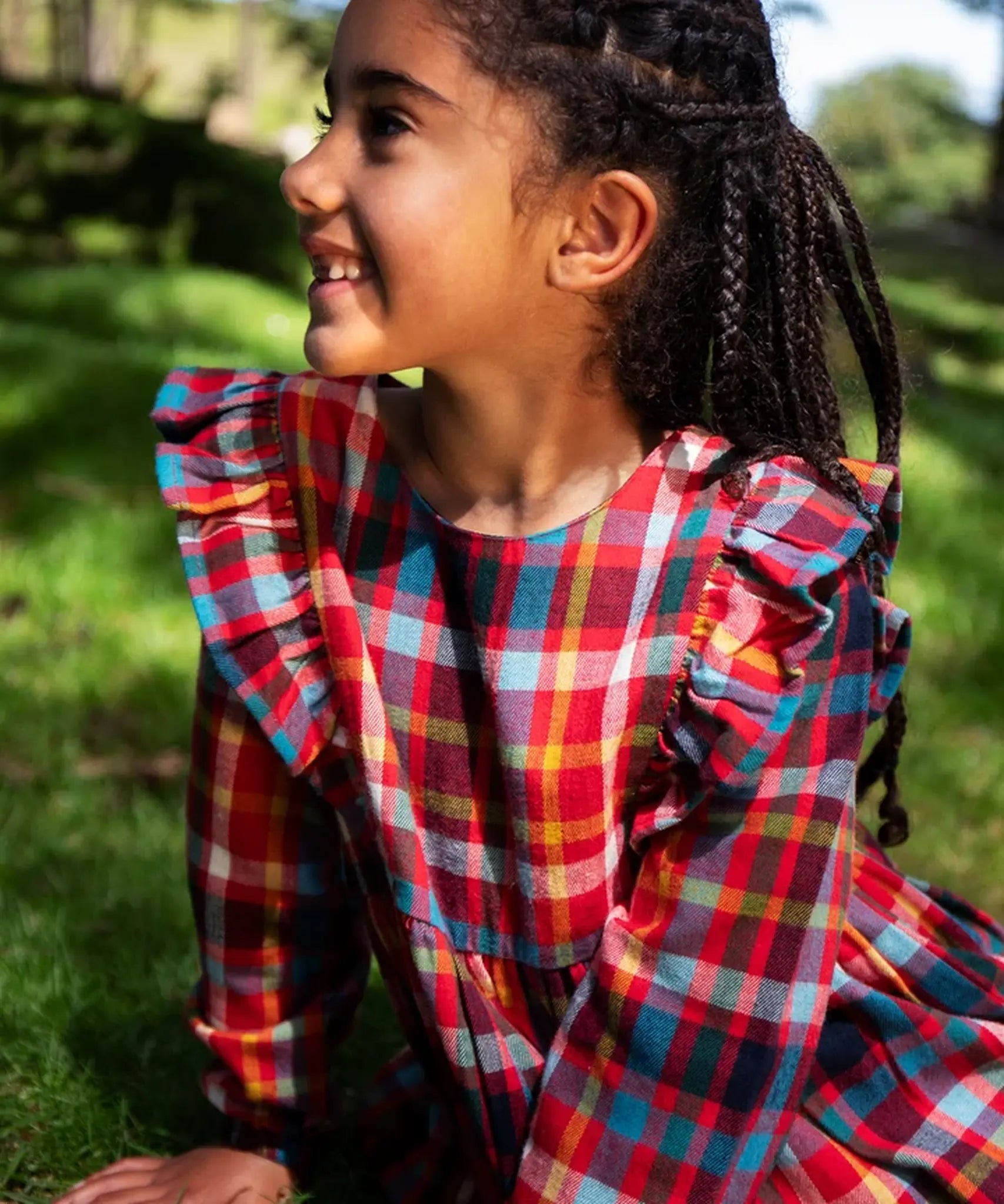 Close-up of a child wearing a Frugi festive check long-sleeve plaid dress outdoors, showing the ruffled shoulder details on the front. The dress features a busy multicolor plaid pattern with red, blue, yellow, green, and white.