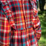 A close-up of a child wearing a Frugi festive check long-sleeve plaid dress outdoors, showing the back of the dress with ruffled shoulder details and a button closure. The dress features a busy multicolor plaid pattern with red, blue, yellow, green, and white.
