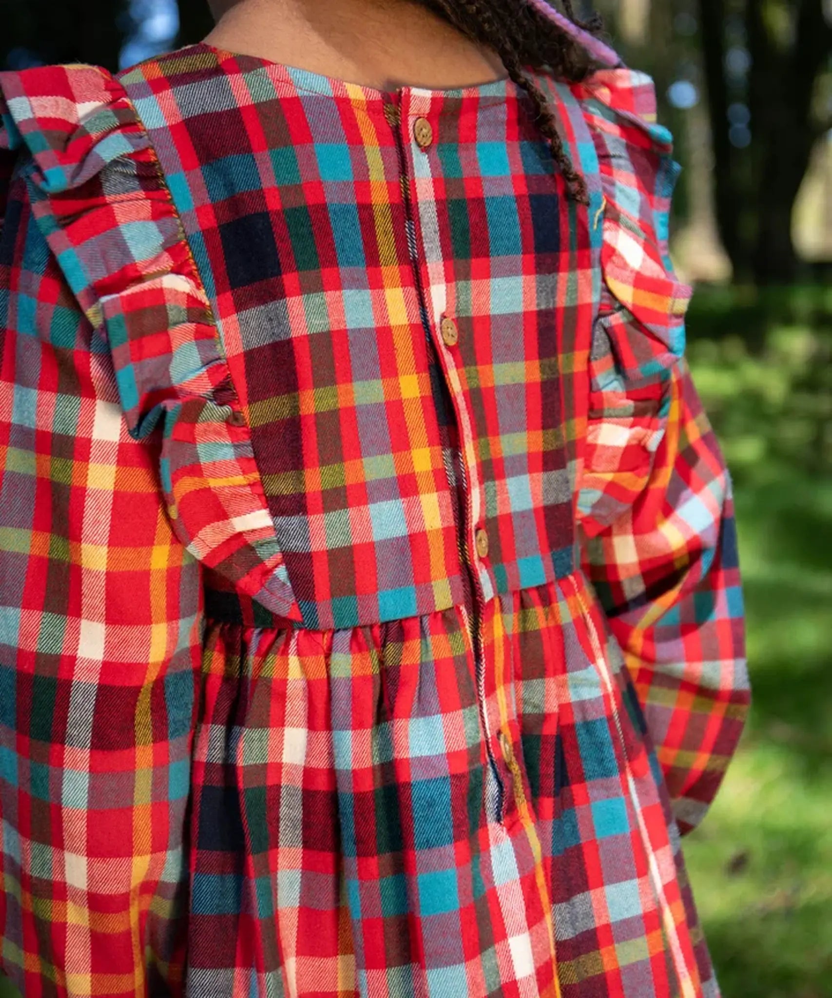 A close-up of a child wearing a Frugi festive check long-sleeve plaid dress outdoors, showing the back of the dress with ruffled shoulder details and a button closure. The dress features a busy multicolor plaid pattern with red, blue, yellow, green, and white.