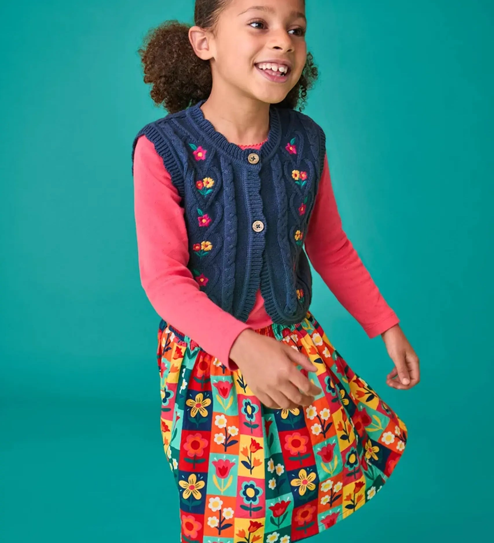 Child wearing the Frugi organic cotton connie navy knitted waistcoat with embroidered flower details, wooden button closure and scalloped edges with a pink long sleeve top underneath and a Marta Twill Skirt with flower stamps design.