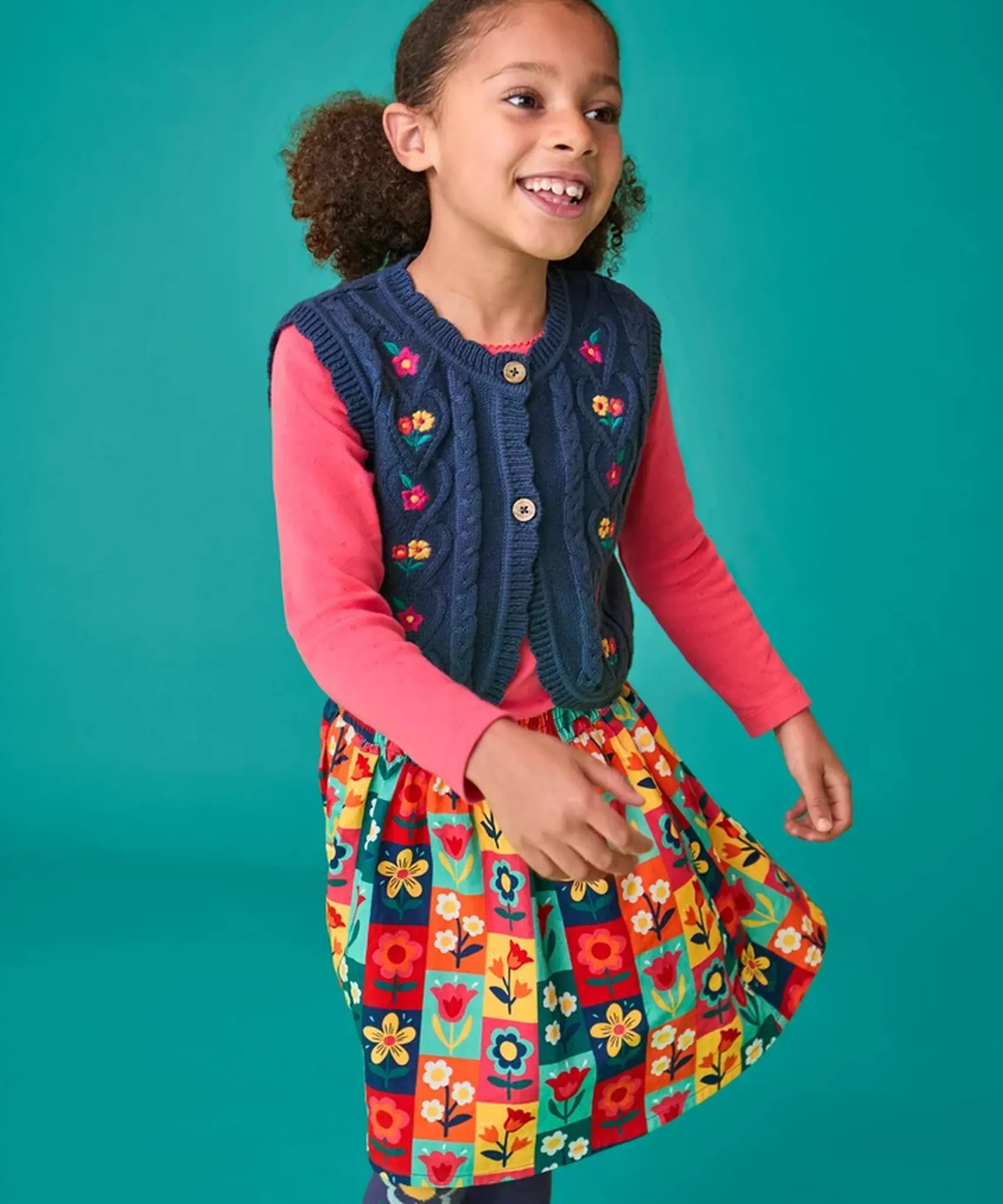 Child wearing the Frugi organic cotton connie navy knitted waistcoat with embroidered flower details, wooden button closure and scalloped edges with a pink long sleeve top underneath and a Marta Twill Skirt with flower stamps design.