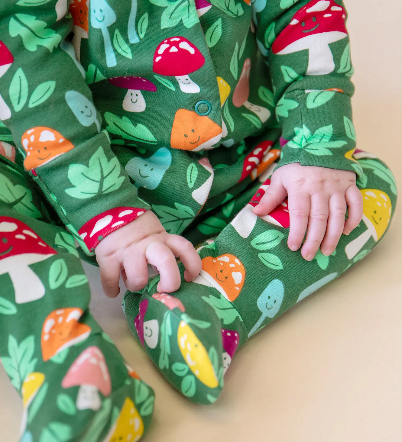 A closeup of a baby's legs and hands, whilst wearing the Frugi organic cotton Lovely Babygrow in a green Fun Guys mushroom print.