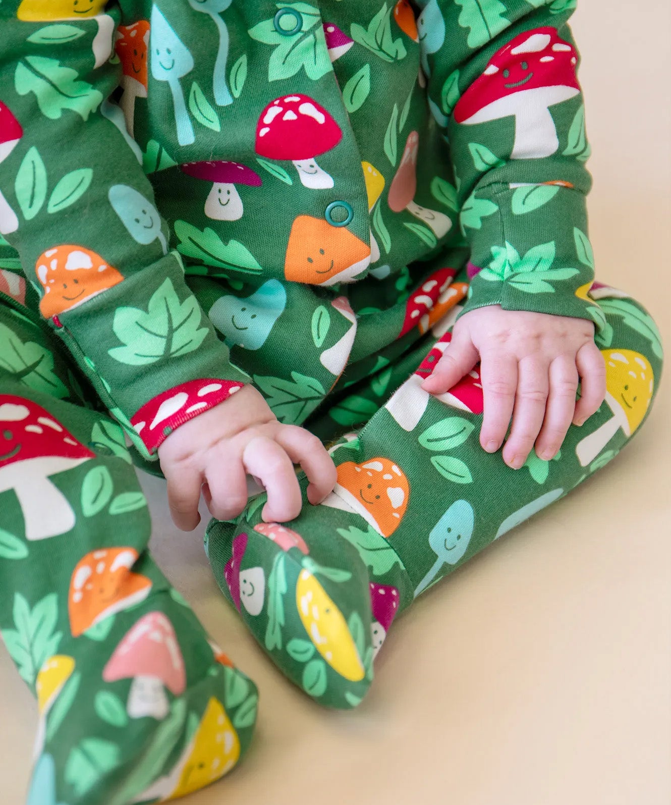 A closeup of a baby's legs and hands, whilst wearing the Frugi organic cotton Lovely Babygrow in a green Fun Guys mushroom print.