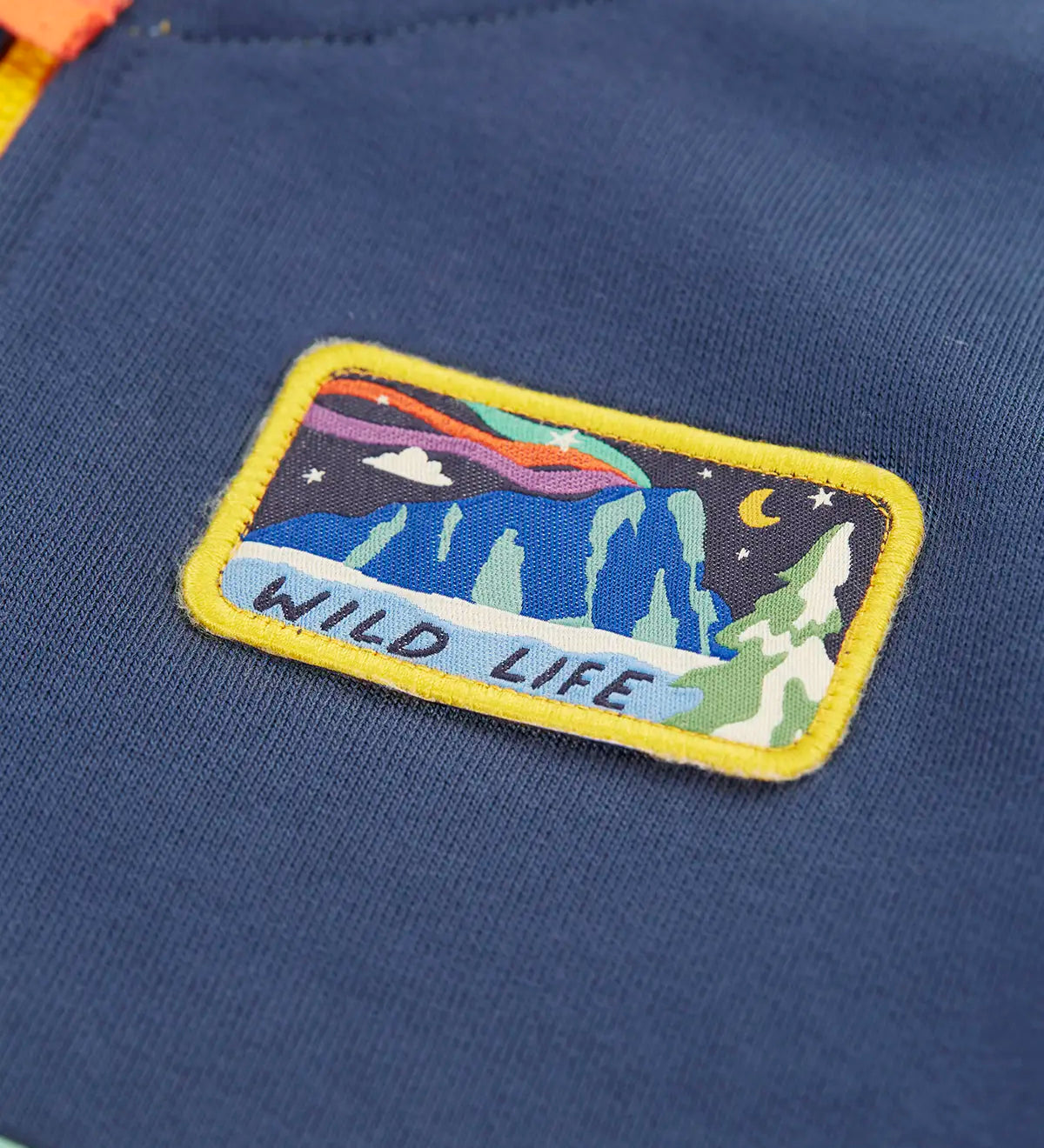 Close up of the rectangle shaped badge on the chest of the Frugi organic cotton half zip sweatshirt hotchpotch wildlife. Yellow edges with a mountainscape with the word wild life in blue underneath.