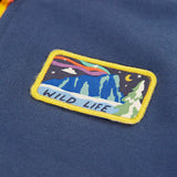 Close up of the rectangle shaped badge on the chest of the Frugi organic cotton half zip sweatshirt hotchpotch wildlife. Yellow edges with a mountainscape with the word wild life in blue underneath.
