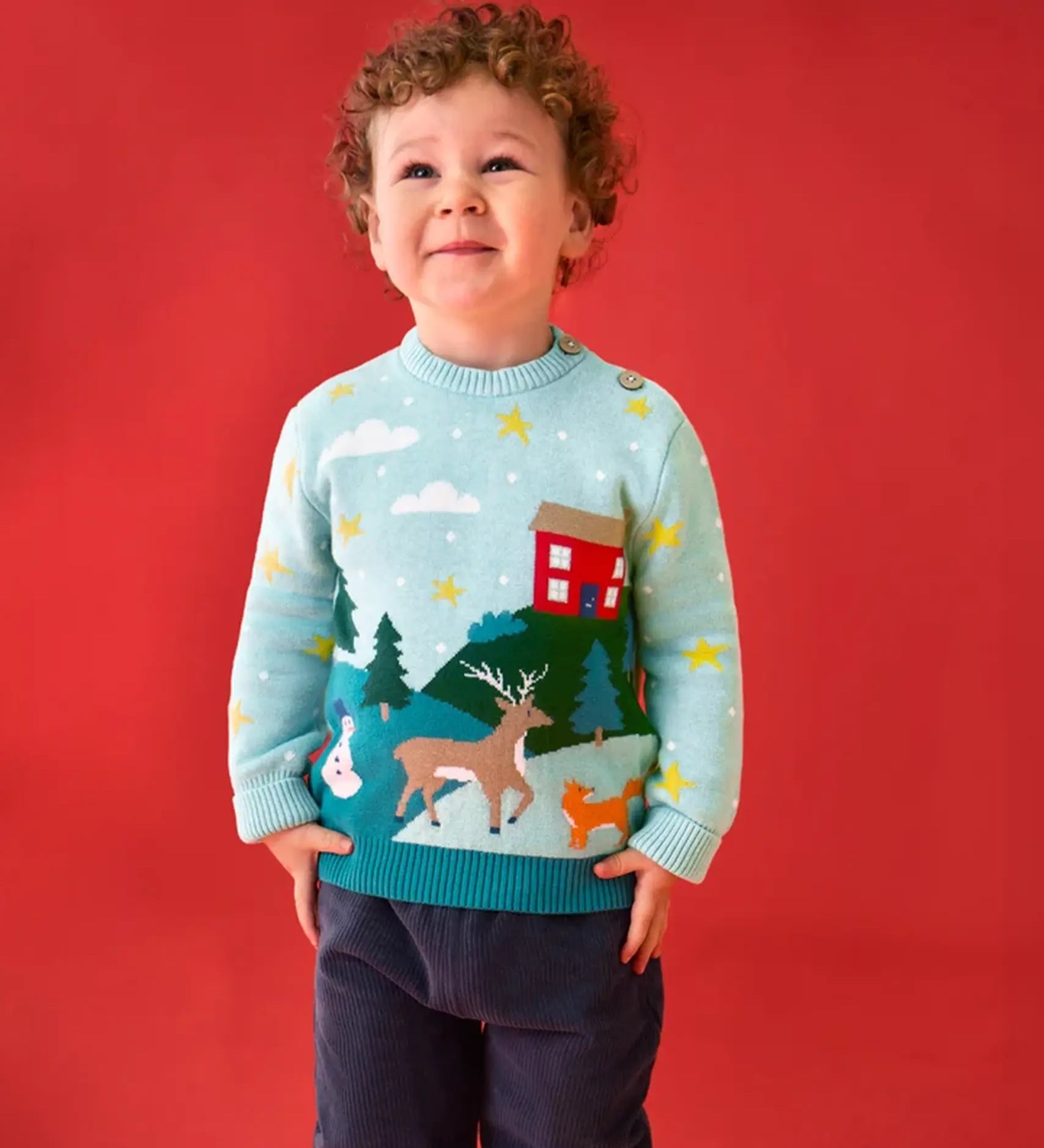 Child wearing the Frugi organic cotton elwood knitted jumper with a Winter tales design. Light blue long sleeved jumper with a landscape design on the front featuring a red house, snowman, reindeer, fox, trees and snow and stars with navy blue corduroy trousers.