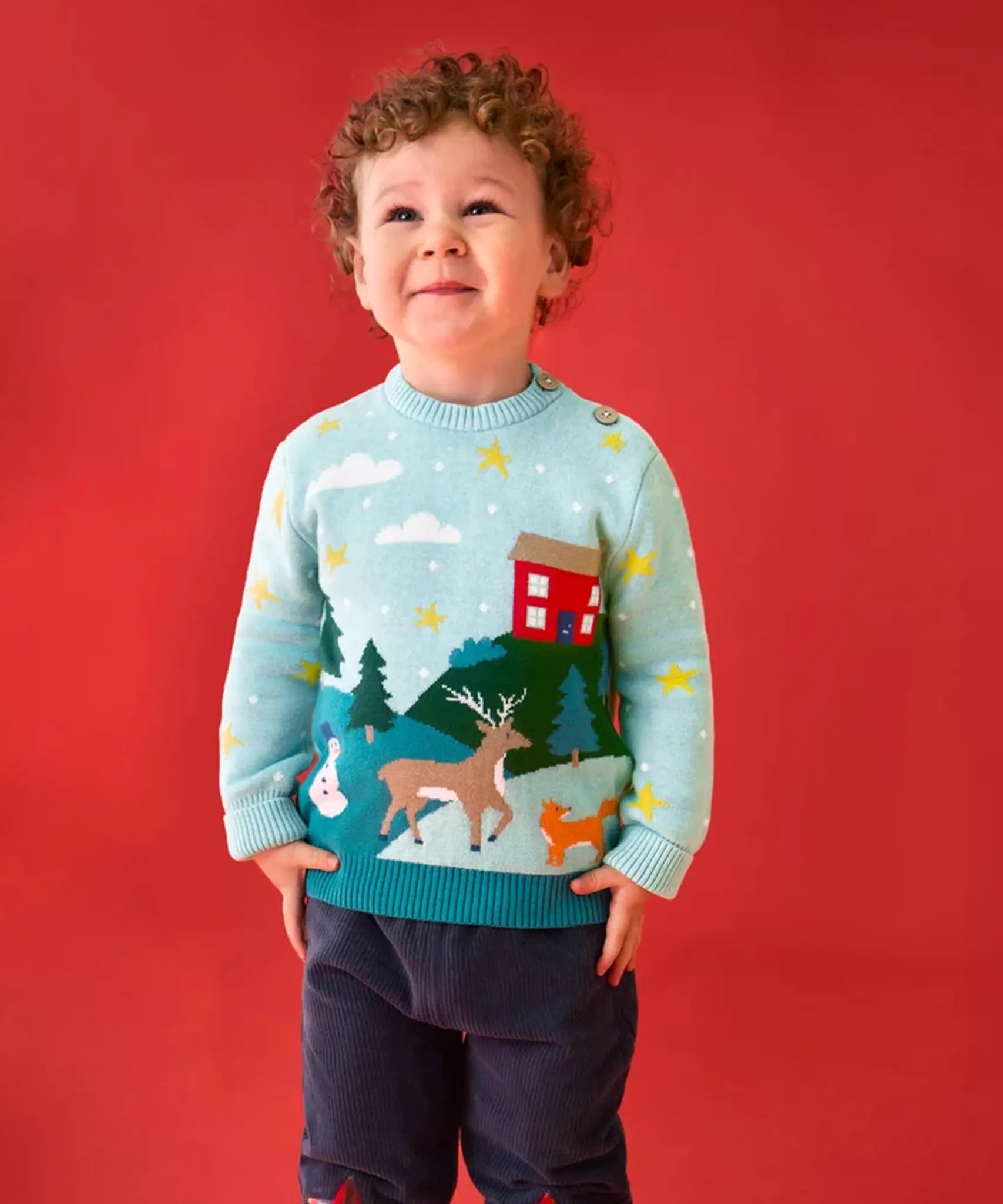 Child wearing the Frugi organic cotton elwood knitted jumper with a Winter tales design. Light blue long sleeved jumper with a landscape design on the front featuring a red house, snowman, reindeer, fox, trees and snow and stars with navy blue corduroy trousers.