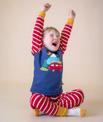 A sleepy child sitting cross legged and stretching his arms ready for bed, wearing the Frugi organic Cotton Jim Jam Pyjamas with a campervan applique design on the front. 