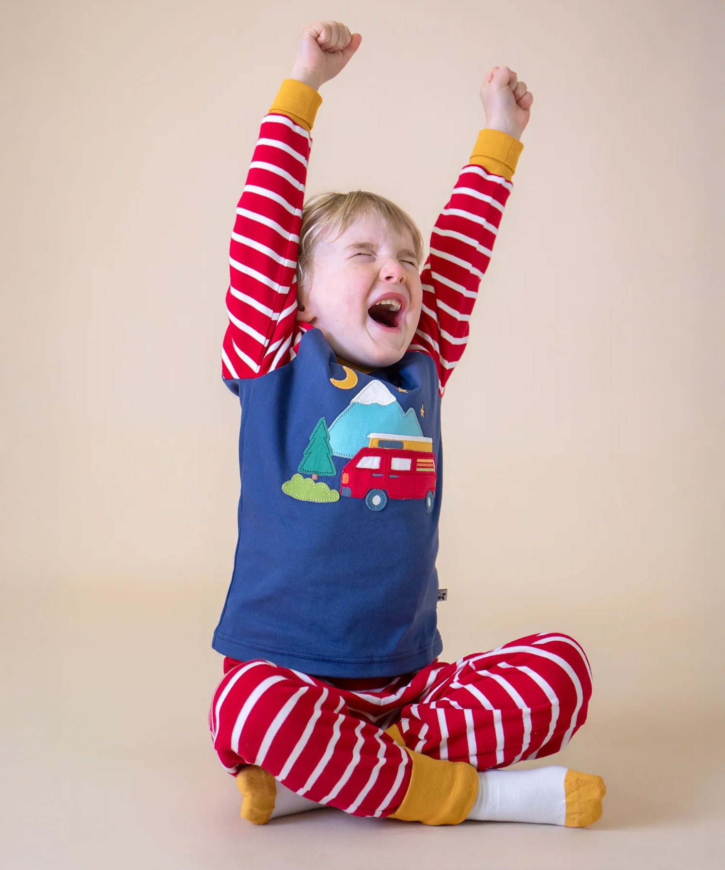 A sleepy child sitting cross legged and stretching his arms ready for bed, wearing the Frugi organic Cotton Jim Jam Pyjamas with a campervan applique design on the front. 