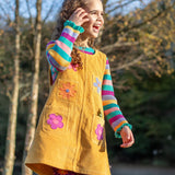 A girl wearing the Frugi organic cotton Zoe Knitted Sweater in a colourful rainbow stripe, with a corduroy yellow pinafore dress layered over top for a cute autumn outfit.