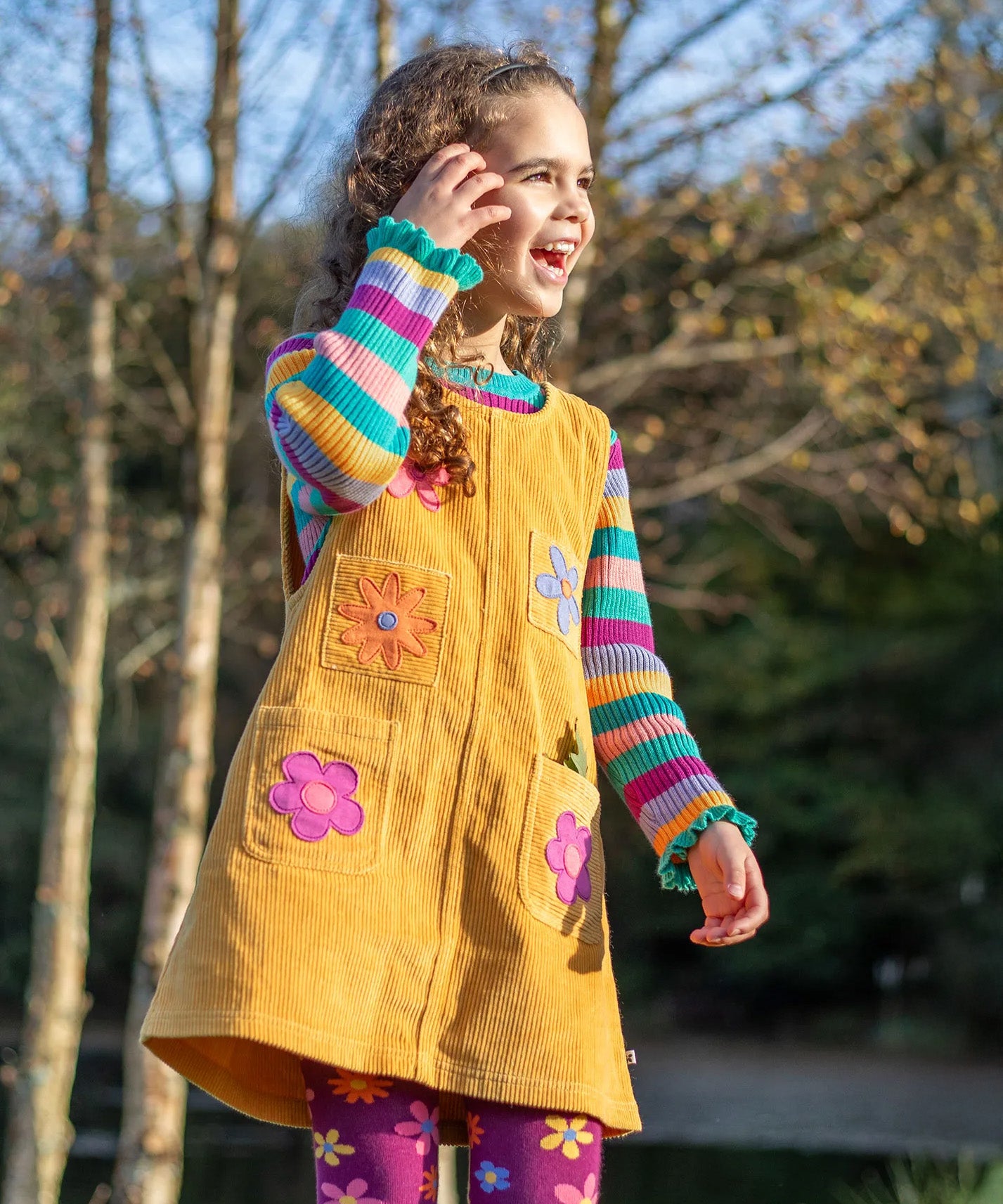 A girl wearing the Frugi organic cotton Zoe Knitted Sweater in a colourful rainbow stripe, with a corduroy yellow pinafore dress layered over top for a cute autumn outfit.