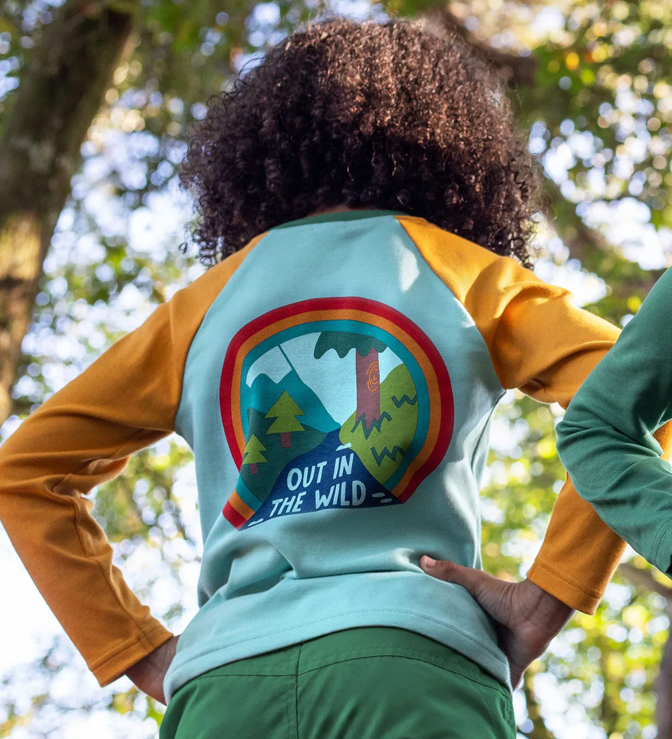 The back of a child in the woods, wearing the Frugi organic cotton long sleeved raglan top for kids, with an 'Out in the Wild' slogan and a colourful graphic on the back.