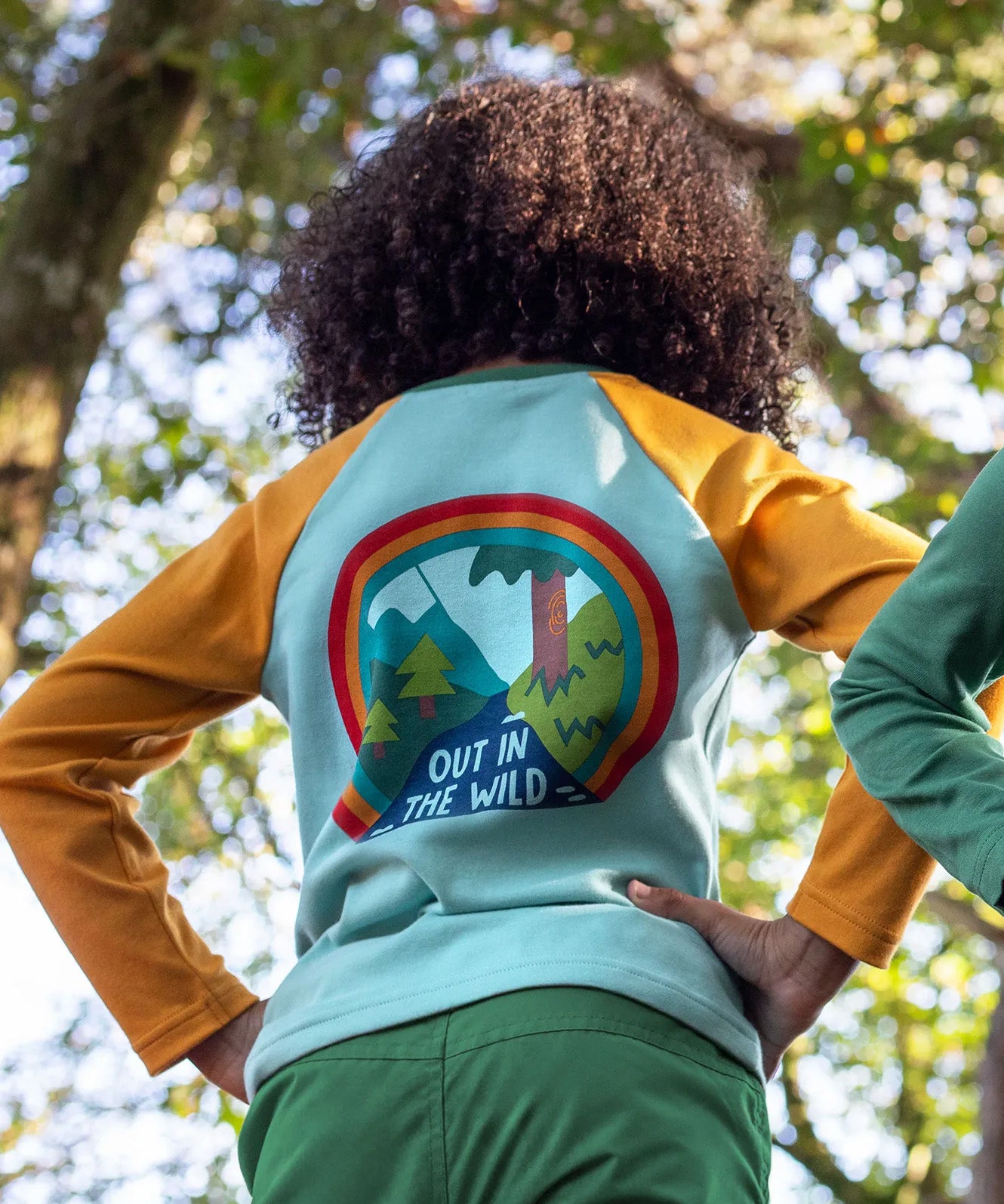 The back of a child in the woods, wearing the Frugi organic cotton long sleeved raglan top for kids, with an 'Out in the Wild' slogan and a colourful graphic on the back.