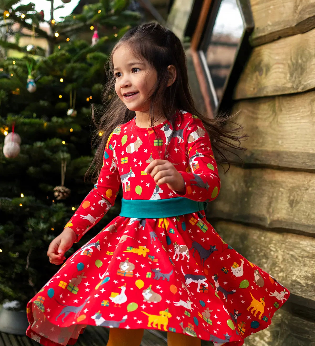 Child twirling wearing the Frugi organic cotton pink party skater dress in a pet paw-ty print featuring pets wearing party hats surrounded by wrapped gifts and balloons. 