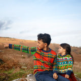 Children sitting outside, one is wearing the Frugi Elwood knitted jumper with a tartan design and the other wearing the Frugi Robyn fairisle cardigan with colourful farm design featuring tractors, highland coos and puffins. Button up front and navy cuffs, neck and waist band. 