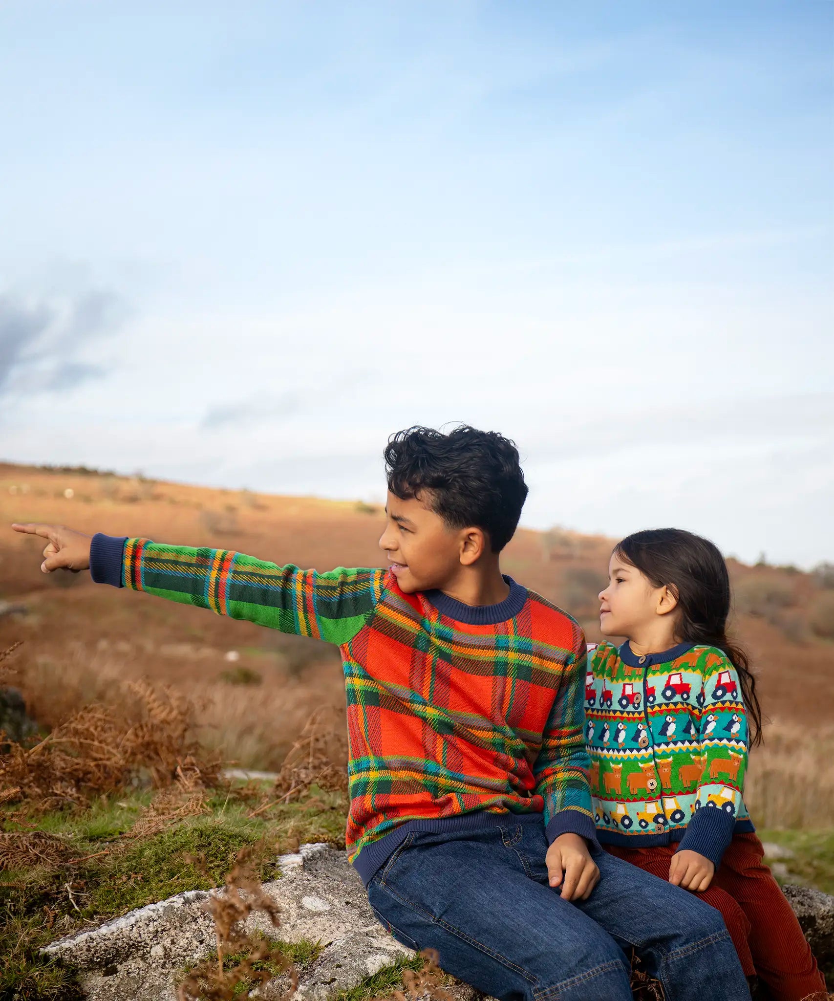 Children sitting outside, one is wearing the Frugi Elwood knitted jumper with a tartan design and the other wearing the Frugi Robyn fairisle cardigan with colourful farm design featuring tractors, highland coos and puffins. Button up front and navy cuffs, neck and waist band. 