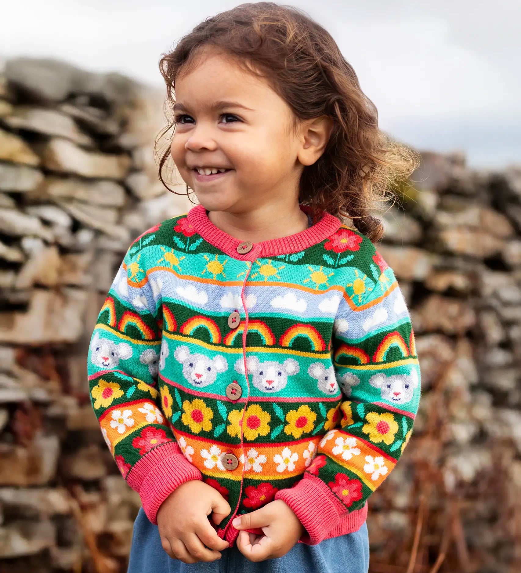 Child wearing the Frugi Robyn fairisle cardigan with a mouse Fair Isle design featuring flowers, rainbows, clouds and suns. 