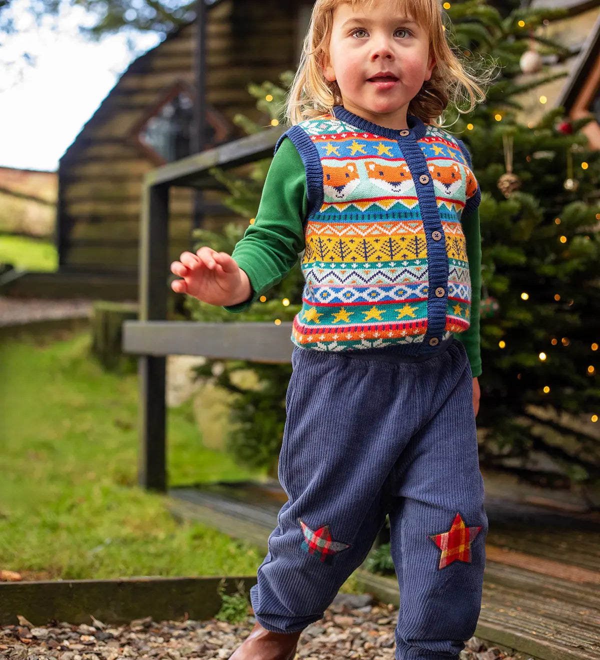 Child wearing the Frugi organic cotton ronnie reversible pull up trousers on the navy cord with festive check star knee patches side with a fairisle style knitted waistcoat and green long sleeved top underneath.