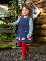 Child wearing the Frugi organic cotton rosalie reversible dress on the side in navy corduroy with festive appliques on the hemline with red tights underneath.