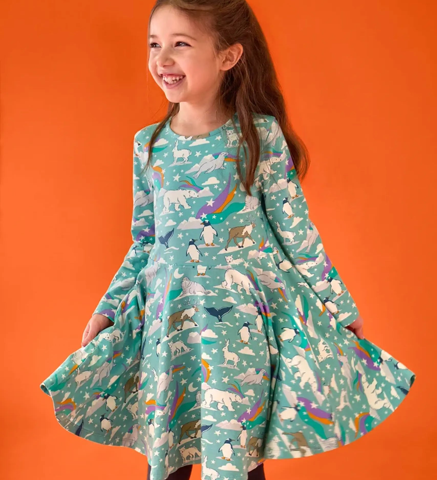 Child wearing the Frugi GOTS organic cotton sofia skater dress in polar aurora. Blue green background with and all over print featuring polar animals among star and colourful swishes. 