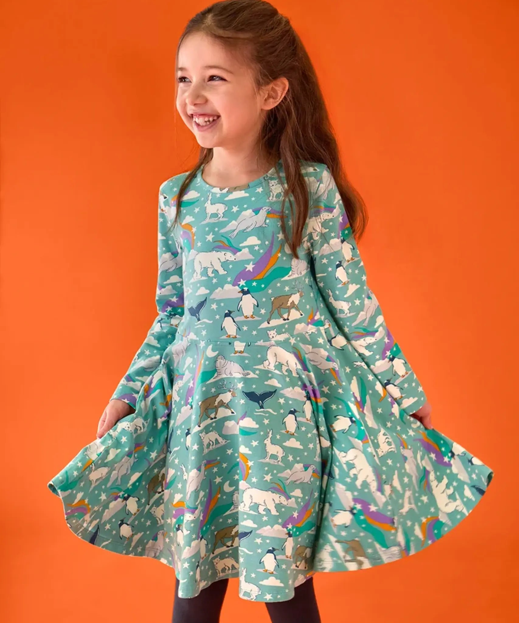 Child wearing the Frugi GOTS organic cotton sofia skater dress in polar aurora. Blue green background with and all over print featuring polar animals among star and colourful swishes. 
