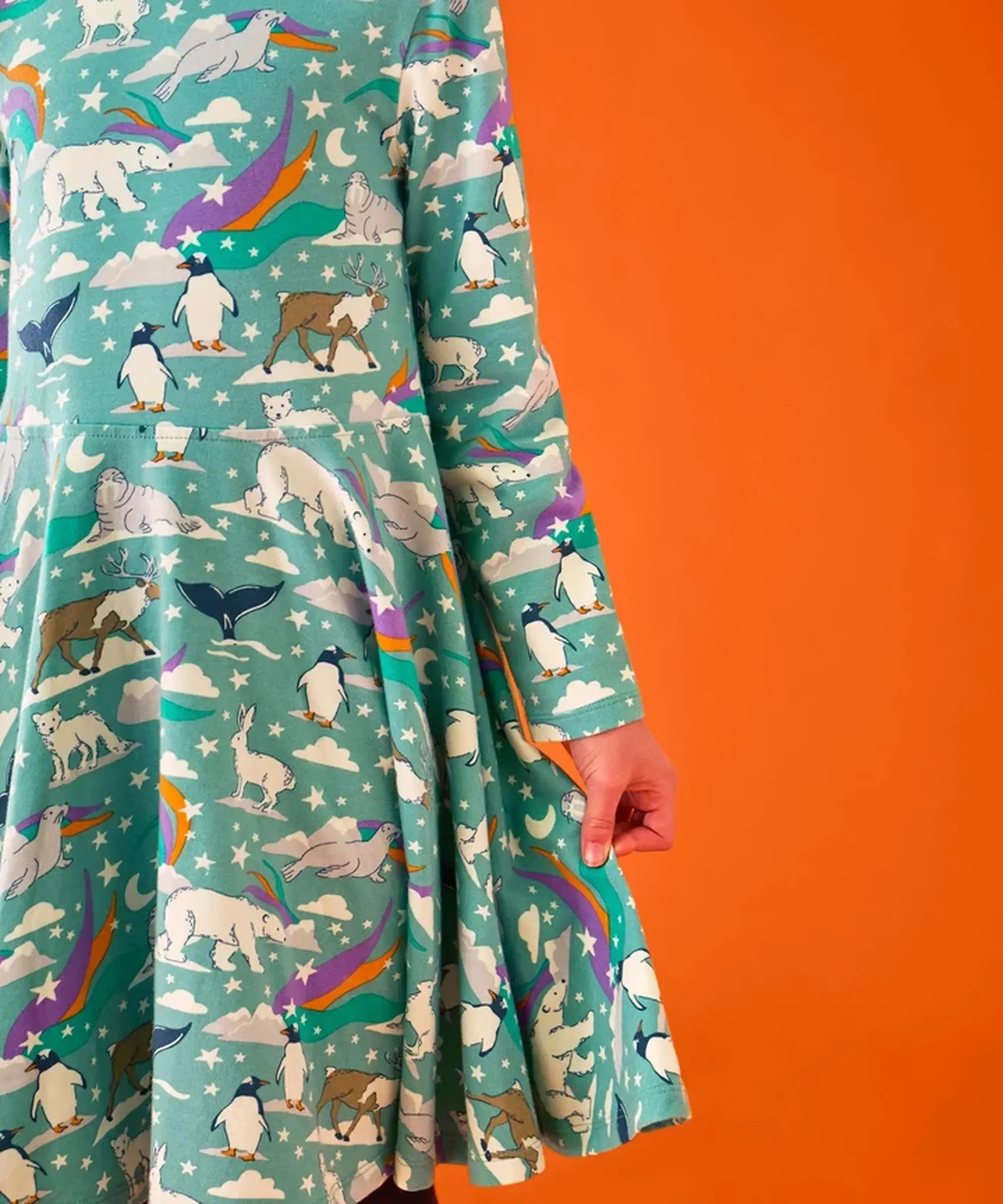 Close up of the side of the Frugi GOTS organic cotton sofia skater dress in polar aurora showing the flared skirt. Blue green background with and all over print featuring polar animals among star and colourful swishes. 