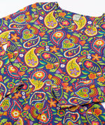 Close up of the details on the Frugi GOTS organic cotton sofia skater dress in pretty paisley. Purple background with colourful paisley print featuring floral and rainbow details.