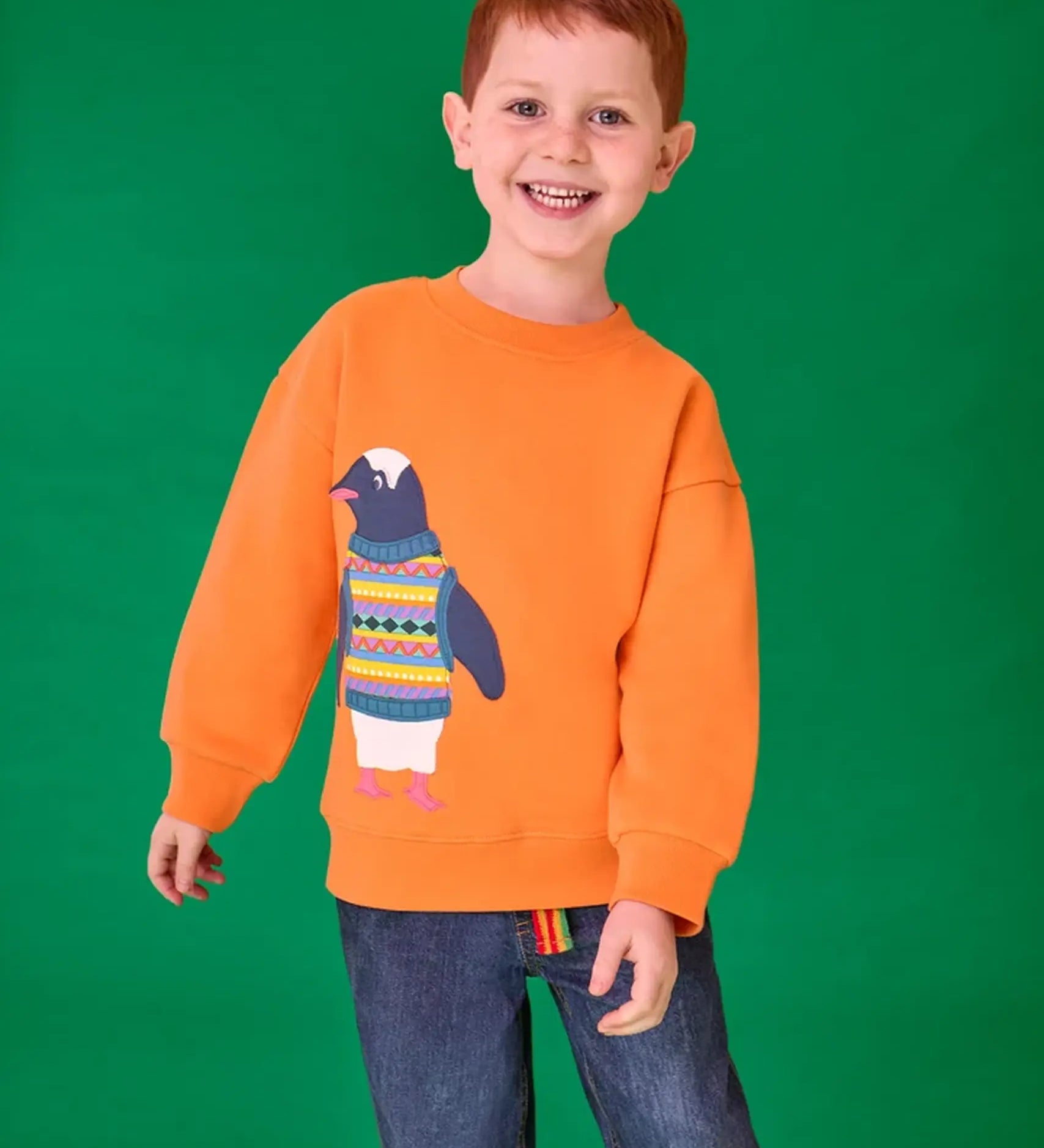 Child wearing the Frugi organic cotton superb sweatshirt salamander orange with penguin applique. The penguin is wearing a fairisle style tank top. 
