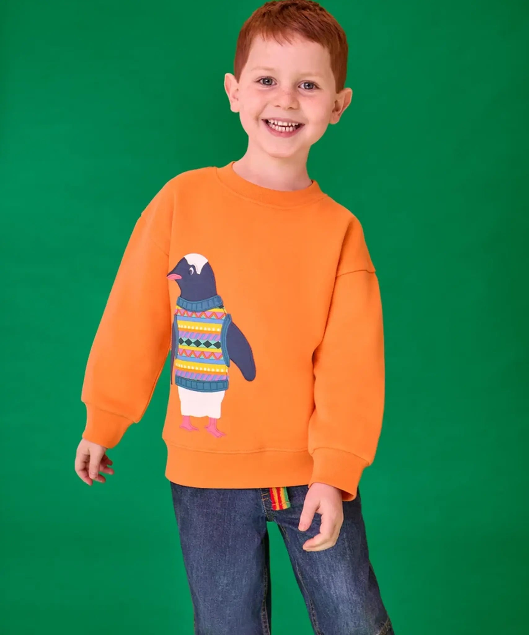 Child wearing the Frugi organic cotton superb sweatshirt salamander orange with penguin applique. The penguin is wearing a fairisle style tank top. 