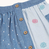 A close up of the wooden buttons down the front of the Frugi denim children's organic denim skirt with side pockets. One side is a darker denim with white polka dots all over with the other side being a lighter denim decorated with embroidered flower outlines.