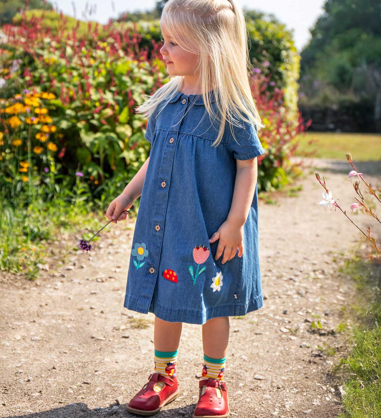 A child with long blonde hair standing on a garden path wearing the Frugi denim dress with button up front and peter pan style collar with flower and ladybug embroidery details along the bottom with red leather t-bar shoes.