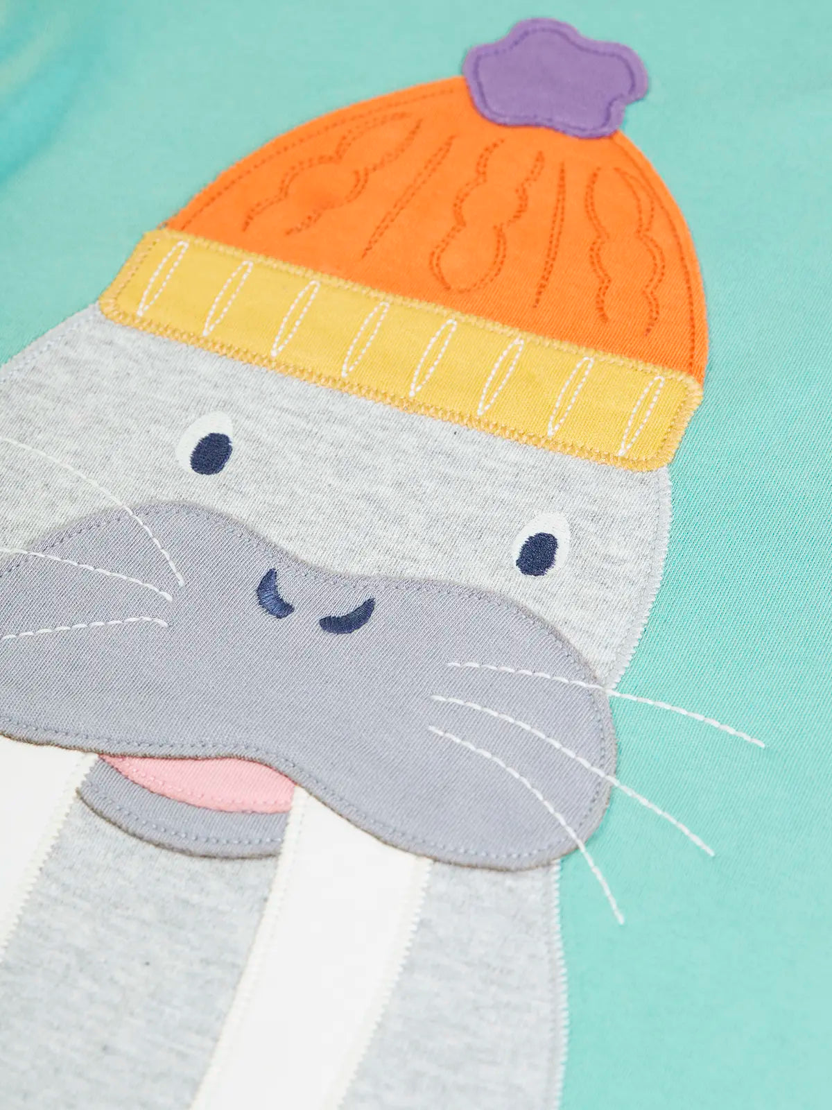 Close up of the large walrus wearing and orange bobble hat applique on the front of the Frugi organic cotton adventure circular long sleeved top in light moss green.
