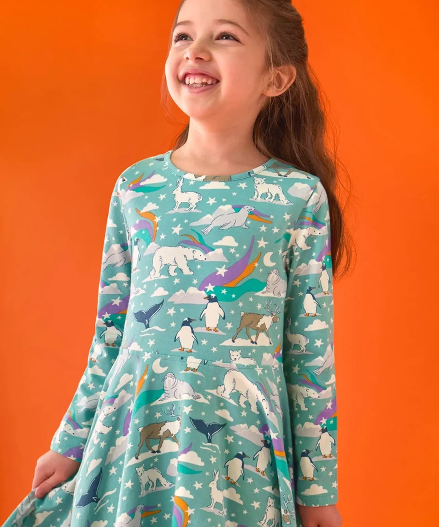 Child wearing the Frugi GOTS organic cotton sofia skater dress in polar aurora. Blue green background with and all over print featuring polar animals among star and colourful swishes. 