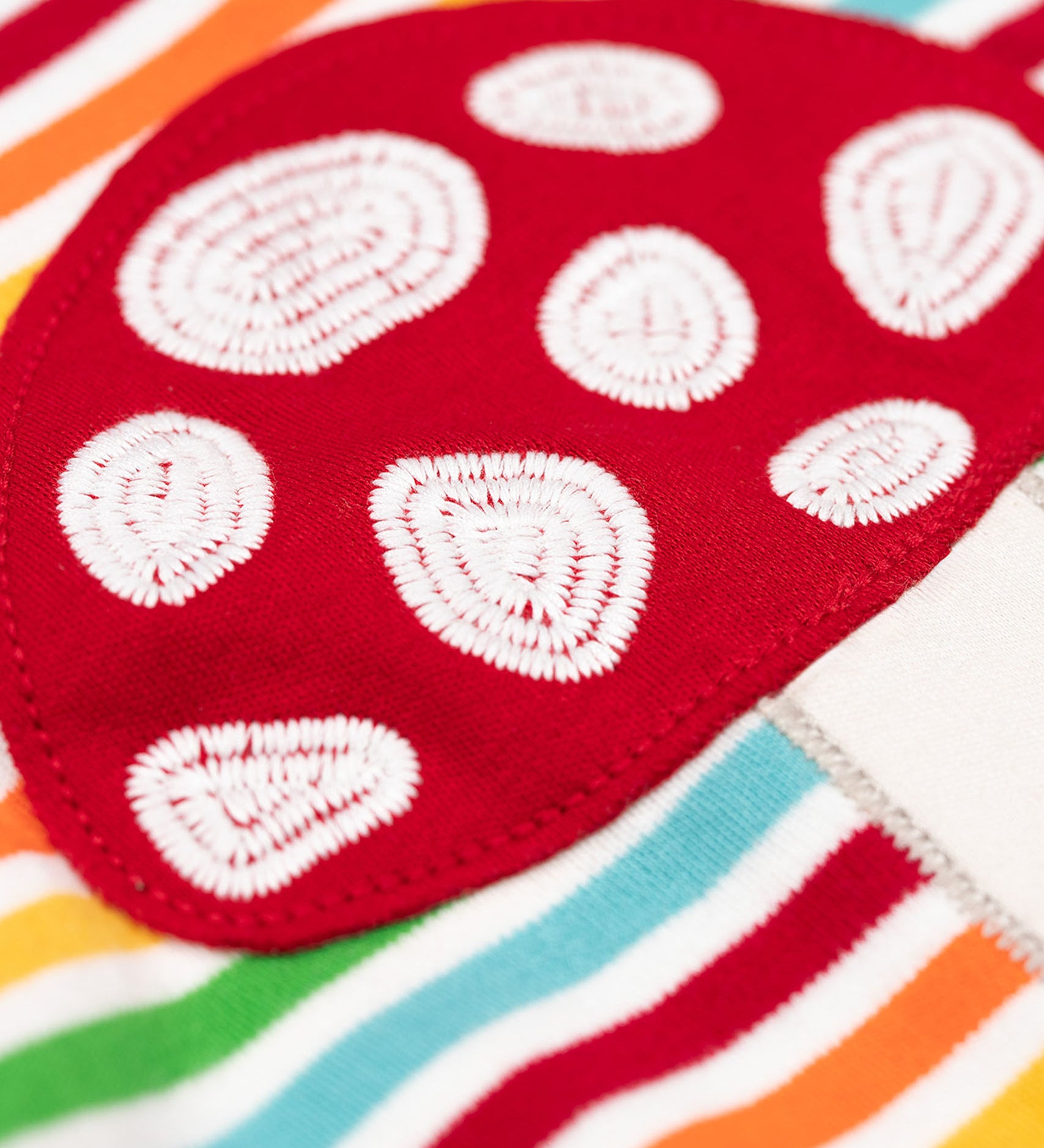 A super close up of the red mushroom applique on the front of the Frugi orion long sleeved, rainbow coloured breton striped child's organic cotton top.