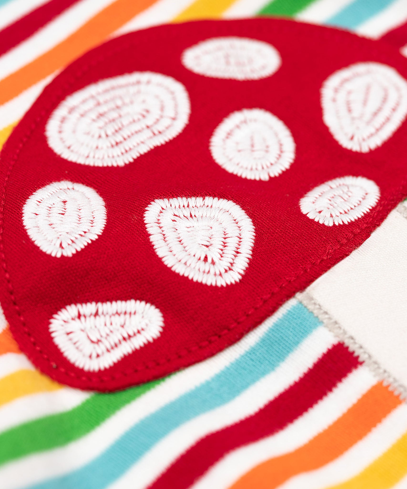 A super close up of the red mushroom applique on the front of the Frugi orion long sleeved, rainbow coloured breton striped child's organic cotton top.