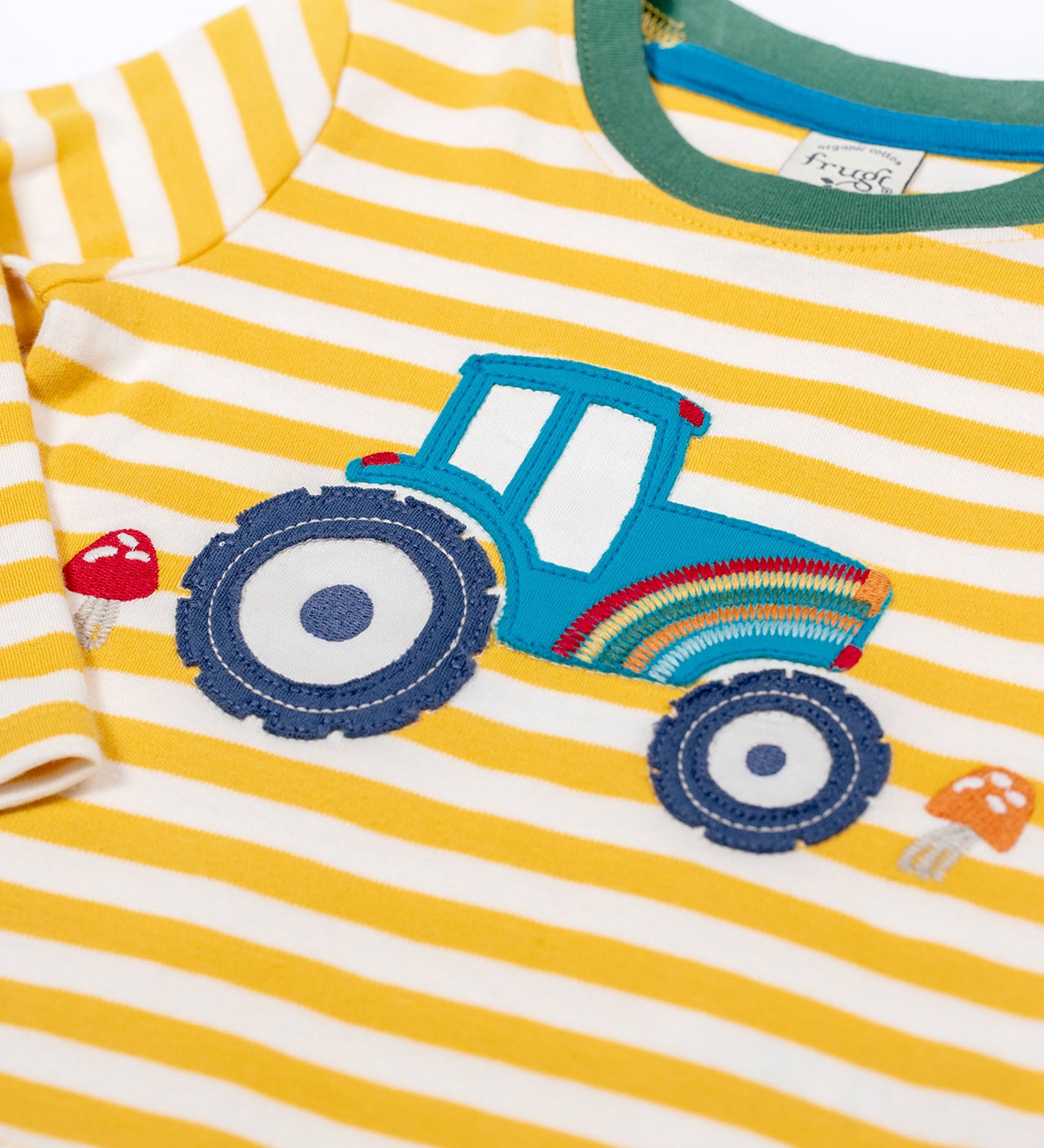 A close up of the tractor applique on the Frugi Orion Bumblebee tractor design long sleeved kids top. The tractor is blue with a rainbow on it's front and two red mushrooms either side of the tractor.