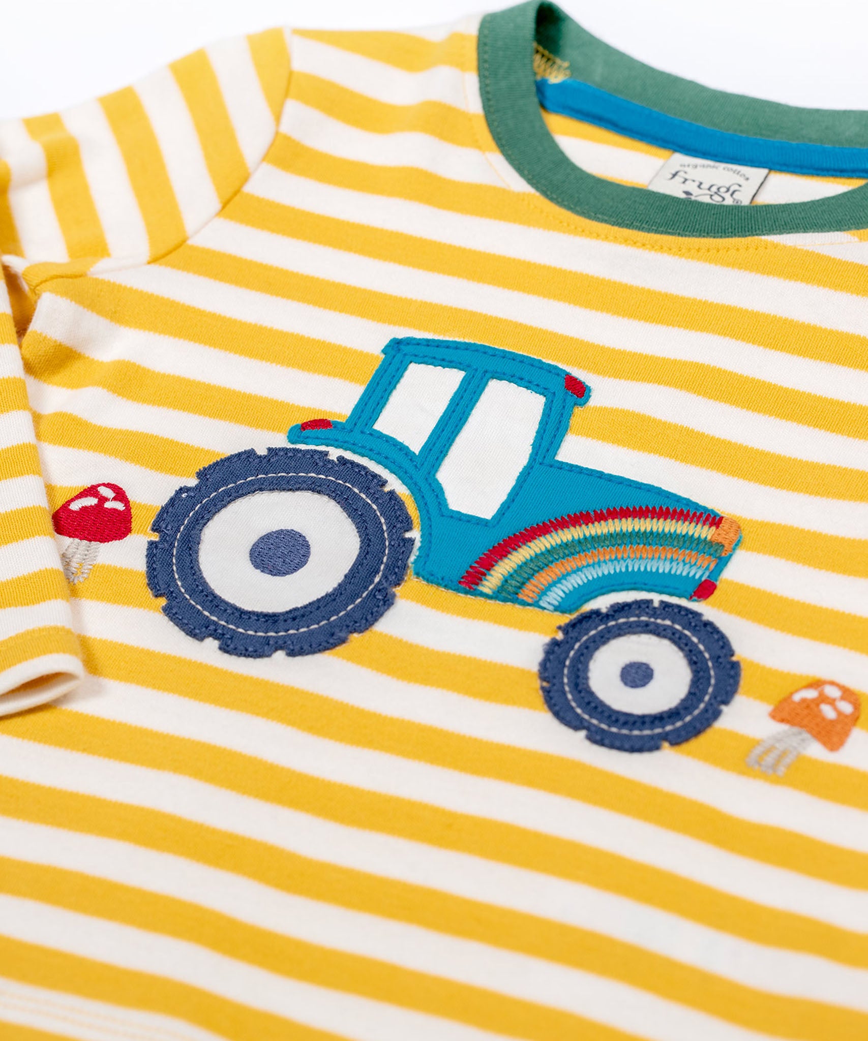 A close up of the tractor applique on the Frugi Orion Bumblebee tractor design long sleeved kids top. The tractor is blue with a rainbow on it's front and two red mushrooms either side of the tractor.