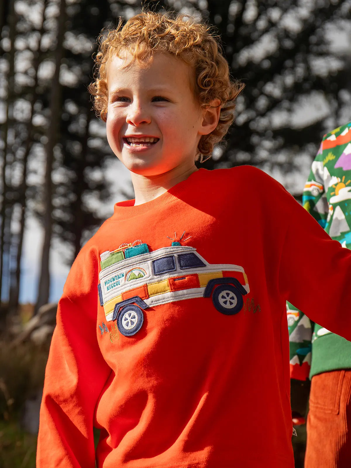Child wearing the Frugi owen organic cotton long sleeved orange with a mountain rescue vehicle on the front. 