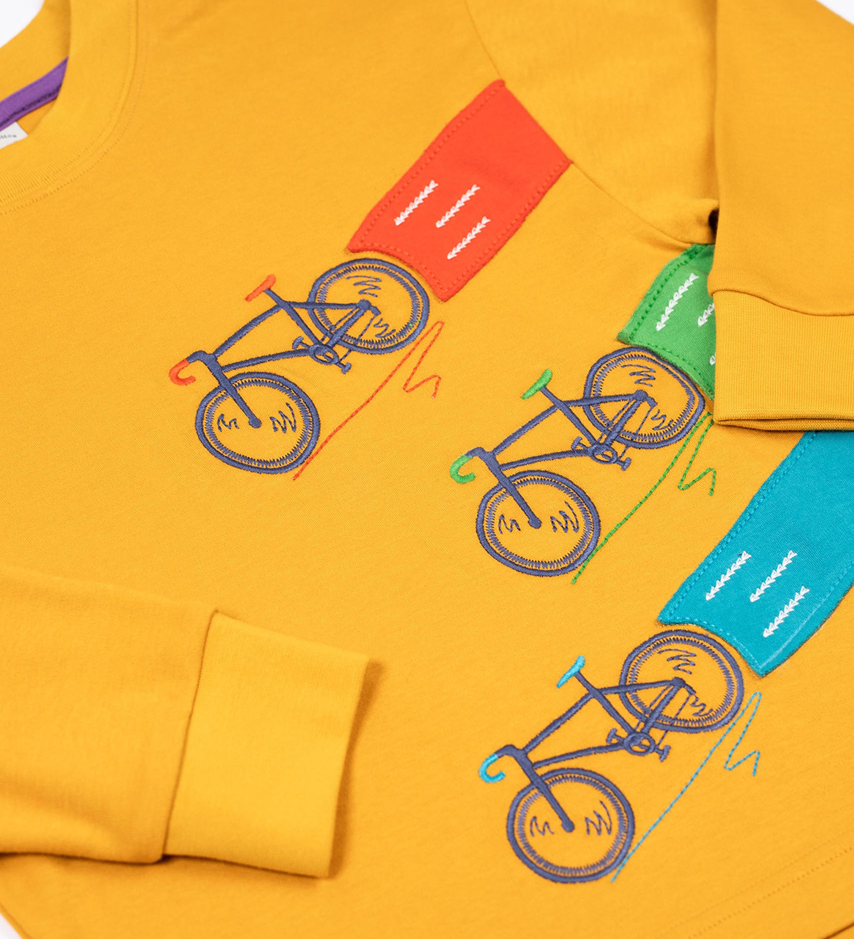 A close up of the embroidered and applique details on the front of the Frugi Owen top.  Three embroidered bicycles each with different coloured applique roads behind them.