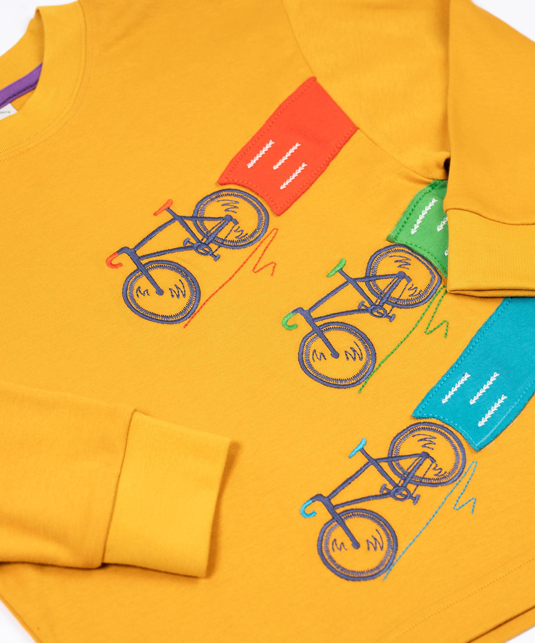 A close up of the embroidered and applique details on the front of the Frugi Owen top.  Three embroidered bicycles each with different coloured applique roads behind them.