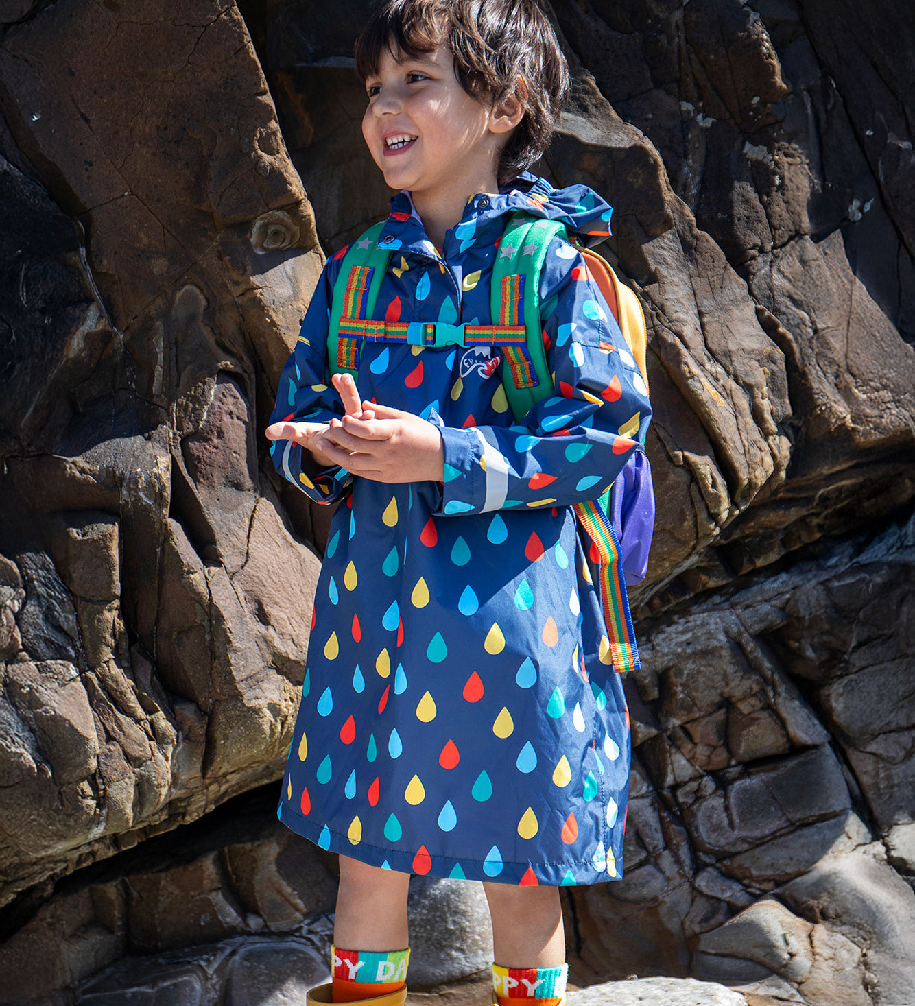 A child smiling, standing in front of rocks on a beach wearing a Frugi pack away over the head navy children's poncho with an all over rainbow rain drop print. The child is wearing a backpack and yellow wellies with colourful socks underneath. 