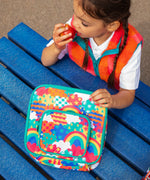 Child eating an apple, holding a Frugi fun frenzy pack a lunch bag. A multicoloured abstract cloud design. Main and a small front compartment and a carry handle.