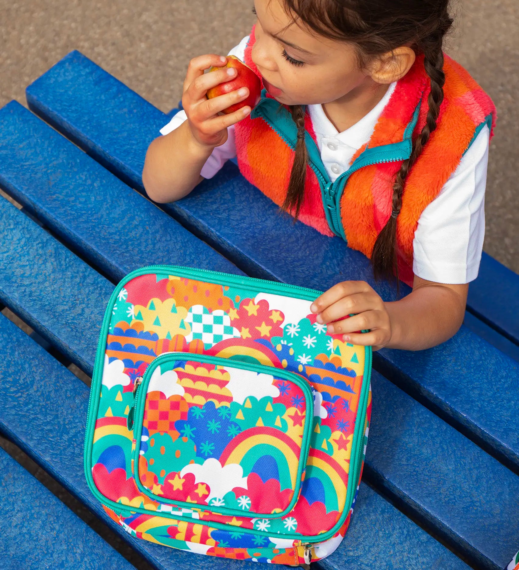 Child eating an apple, holding a Frugi fun frenzy pack a lunch bag. A multicoloured abstract cloud design. Main and a small front compartment and a carry handle.